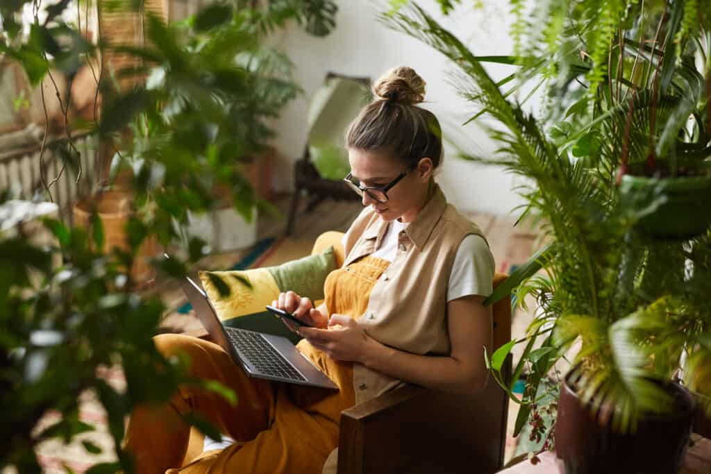 Young florist in eyeglasses sitting on armchair with laptop computer on her knees and using mobile phone in the room with plants